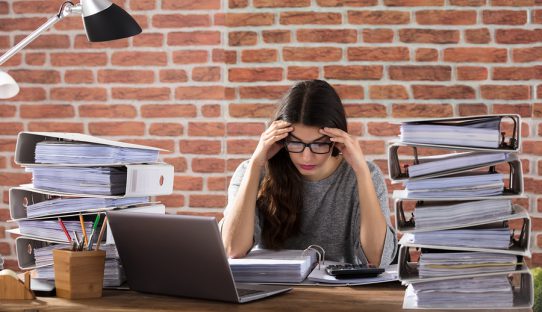 Exhausted Businesswoman Working At Office With Stack Of Folders On Desk